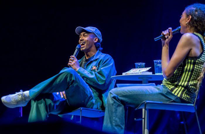 Two people sit on stage under blue lighting, holding microphones. Singer, Jordan Stephens is on the left, in a cap and blue outfit and appears to be laughing. The atmosphere is lively.