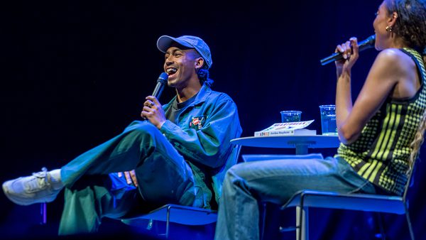 Two people sit on stage under blue lighting, holding microphones. Singer, Jordan Stephens is on the left, in a cap and blue outfit and appears to be laughing. The atmosphere is lively.