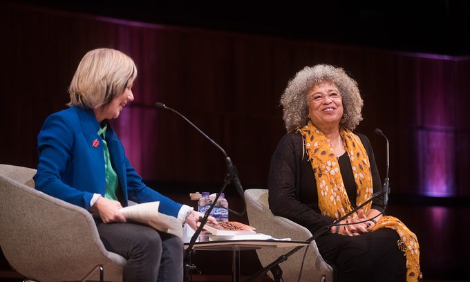 Two women are seated on stage in conversation. One wears a blue blazer, the other a yellow scarf. Both appear engaged, with a warm atmosphere.