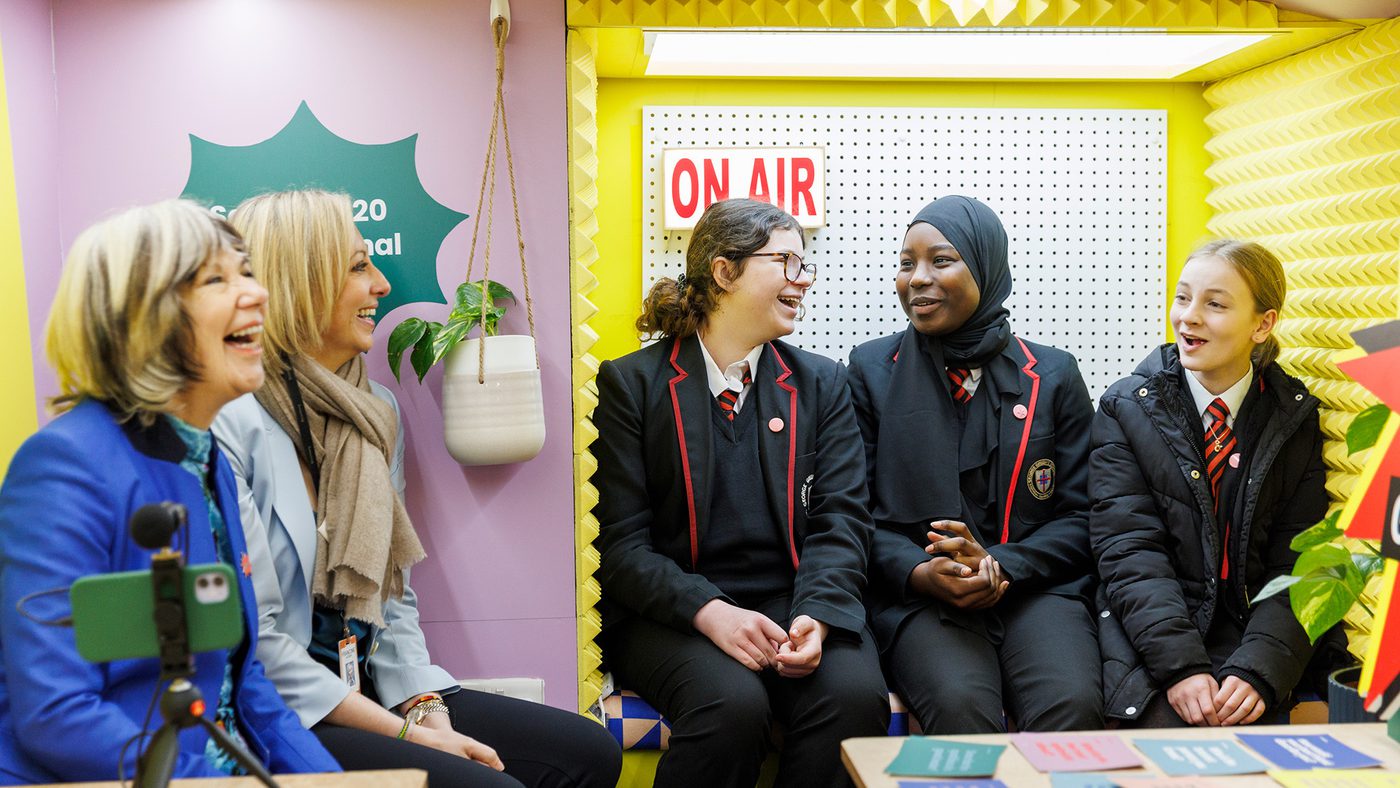A group of people, including adults and schoolgirls in uniforms, sit in a colourful room with "On Air" sign, smiling and laughing, creating a cheerful atmosphere.