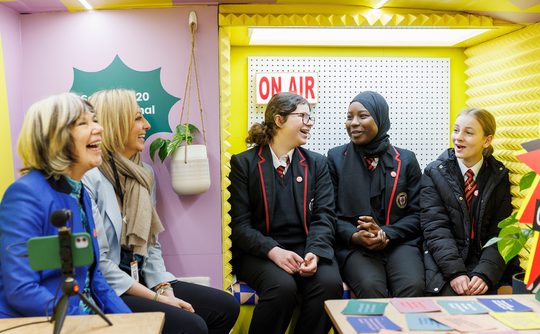 A group of people, including adults and schoolgirls in uniforms, sit in a colourful room with "On Air" sign, smiling and laughing, creating a cheerful atmosphere.