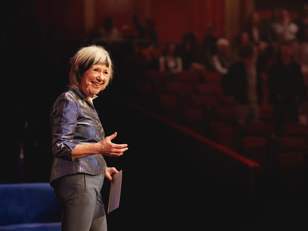 Jude Kelly stands smiling on a stage holding papers. She's in a metallic blouse, addressing a blurred, seated audience in a warm-lit auditorium.