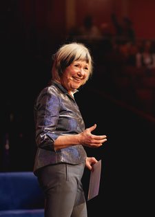 Jude Kelly stands smiling on a stage holding papers. She's in a metallic blouse, addressing a blurred, seated audience in a warm-lit auditorium.