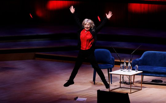 A person in a red shirt joyfully jumps on a stage with a broad smile.