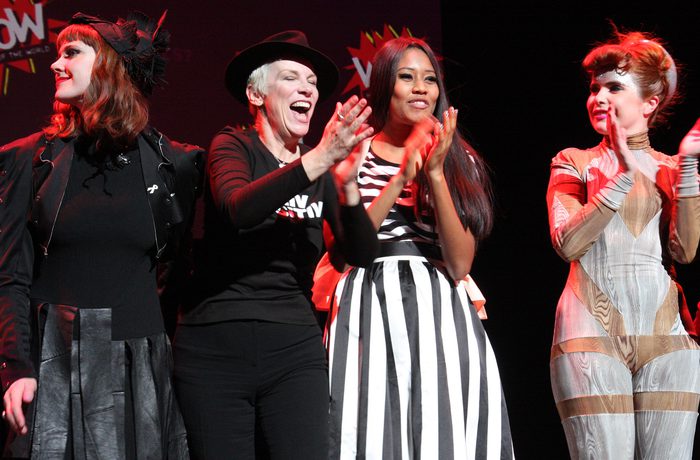 Four women stand on stage clapping, wearing diverse, stylish outfits; the mood is celebratory and joyful.