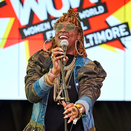 A woman joyfully sings into a microphone on stage, wearing sunglasses and layered jewellry. Behind her, a vibrant banner reads "Women of the World Festival, Baltimore."