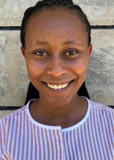 Smiling person with braided hair wearing a striped shirt stands against a textured concrete wall