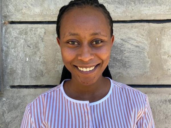 Smiling person with braided hair wearing a striped shirt stands against a textured concrete wall