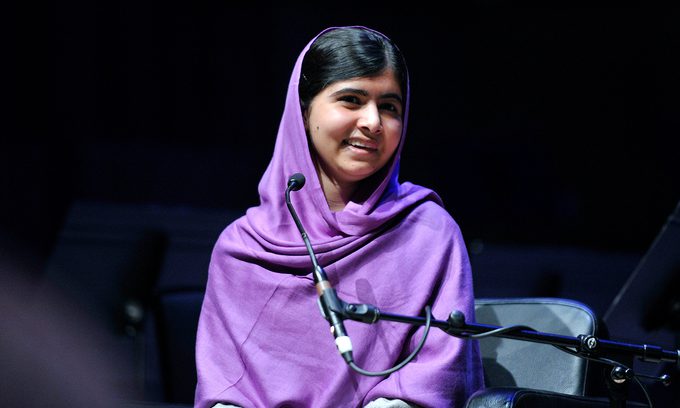 Young woman in a purple headscarf sits on stage, smiling warmly beside a microphone.
