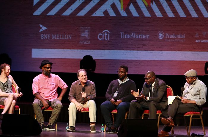Six men sit on red chairs on stage with microphones; the man in the middle is speaking.