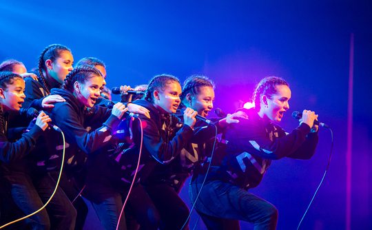 A group of energetic performers in matching black outfits sing passionately on stage, microphones in hand, against a backdrop of vibrant blue and pink lights.