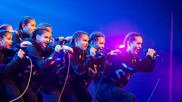 A group of energetic performers in matching black outfits sing passionately on stage, microphones in hand, against a backdrop of vibrant blue and pink lights.