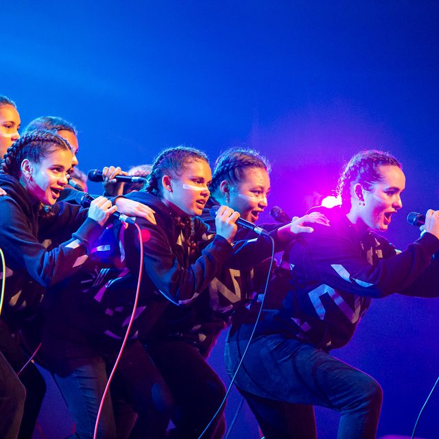 A group of energetic performers in matching black outfits sing passionately on stage, microphones in hand, against a backdrop of vibrant blue and pink lights.