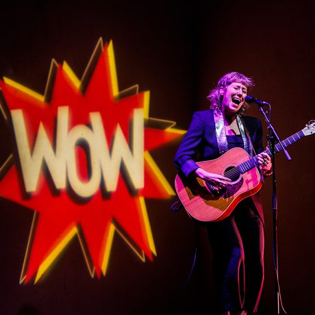 A musician passionately sings and plays guitar onstage, with a large "Wow" logo projected behind them