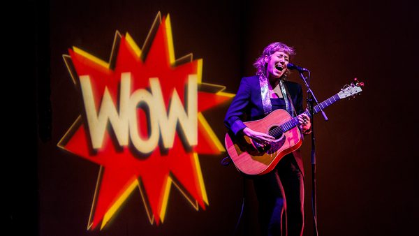 A musician passionately sings and plays guitar onstage, with a large "Wow" logo projected behind them