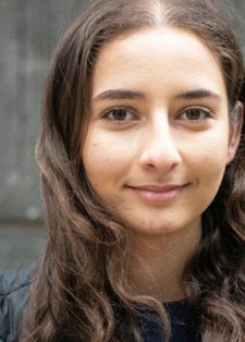 Young woman with long wavy hair, wearing a black jacket, stands against a grey concrete wall smiling subtly