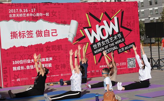 A group of people practice yoga outdoors against a vibrant red "Women of the World Festival" banner. They sit on mats, stretching with raised arms.