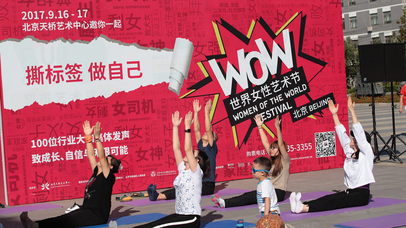 A group of people practice yoga outdoors against a vibrant red "Women of the World Festival" banner. They sit on mats, stretching with raised arms.