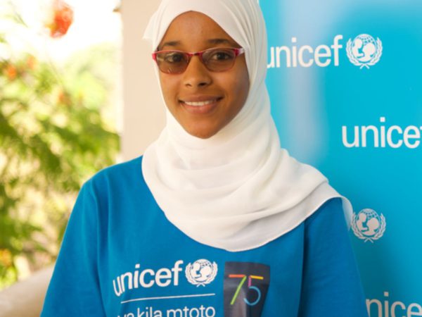 A smiling person wearing a white hijab and red glasses stands in front of a blue UNICEF banner