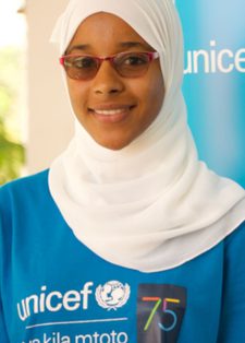A smiling person wearing a white hijab and red glasses stands in front of a blue UNICEF banner