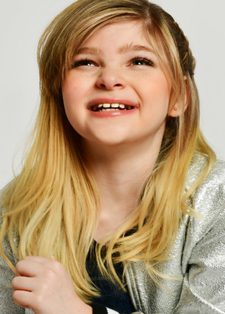 Smiling child with long blond hair, wearing a shiny silver jacket over a black shirt