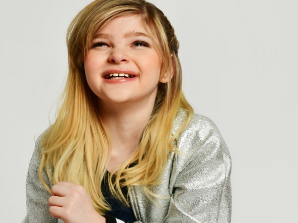 Smiling child with long blond hair, wearing a shiny silver jacket over a black shirt