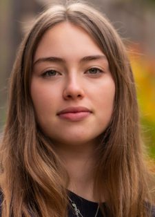 A young woman with long brown hair stands outdoors, looking confidently at the camera