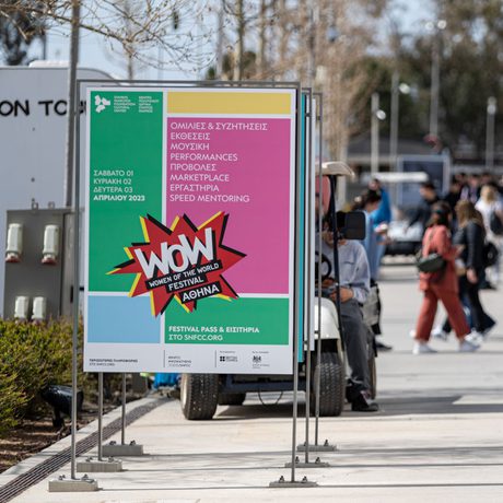Outdoor festival scene showing a colourful sign for "WoW Festival" in the foreground. People casually walk in the sunny background