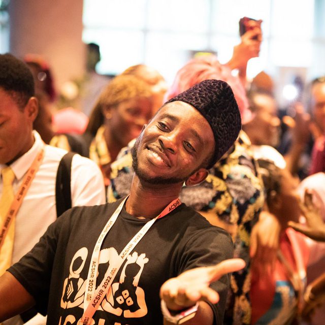 A joyful man in a crowd smiles with open arms, wearing a black T-shirt and a lanyard.