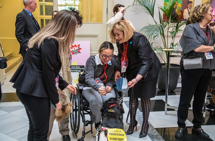 A woman in a wheelchair pets a guide dog, surrounded by smiling people in a bright room with plants.