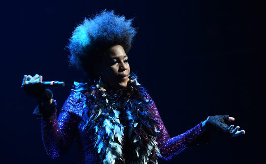 A woman is performing on stage, wearing a sparkly outfit and feather boa. She's looking at the audience and holding microphone.