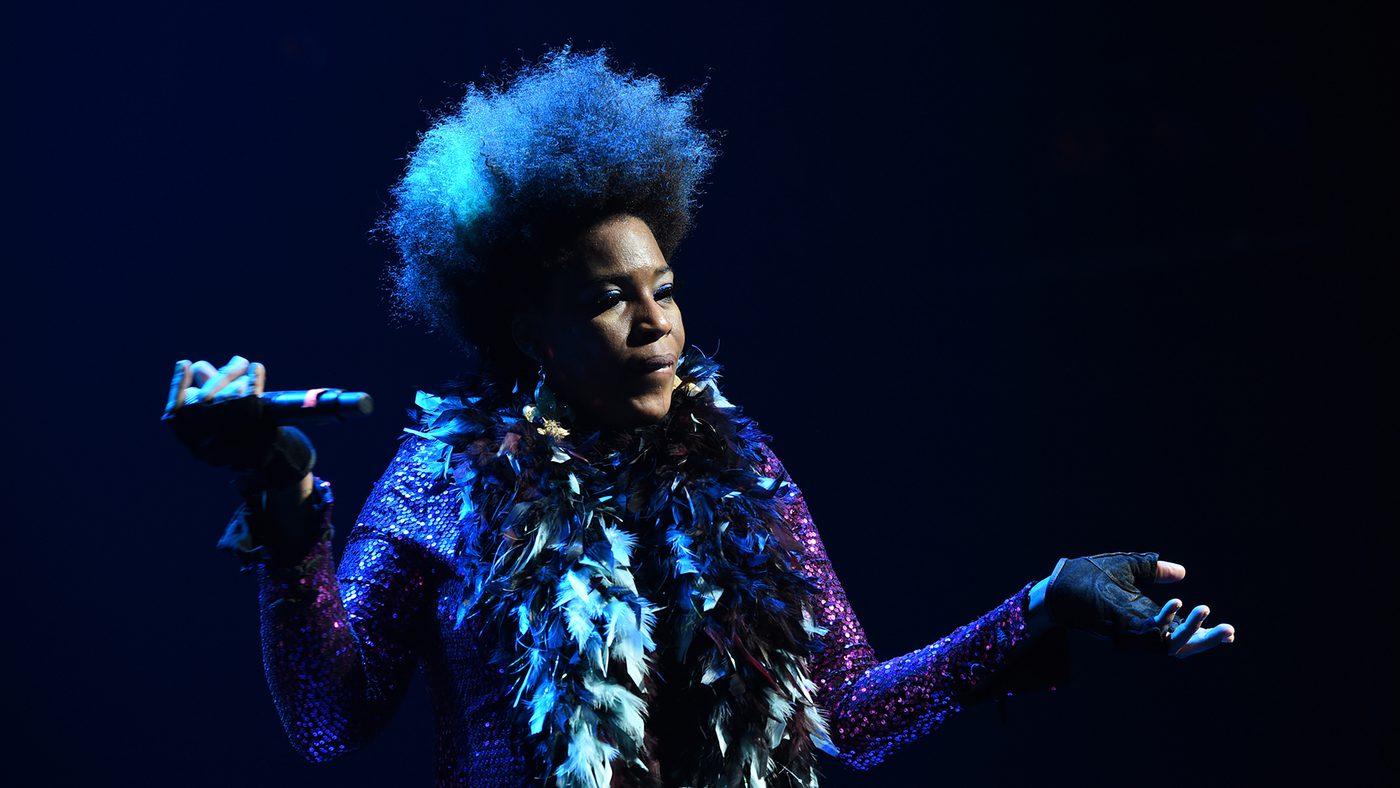 A woman is performing on stage, wearing a sparkly outfit and feather boa. She's looking at the audience and holding microphone.