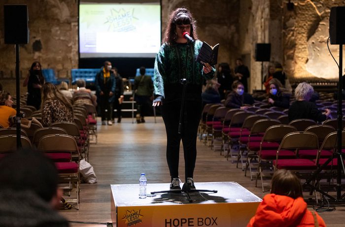 A woman stands on a brown box with the words Hope Box on it in a dark performance space. She is speaking into a microphone and is holding a book in front of her.