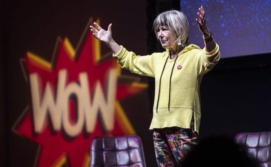 Jude (a white woman in a yellow top) stands on a stage with her hands in the air