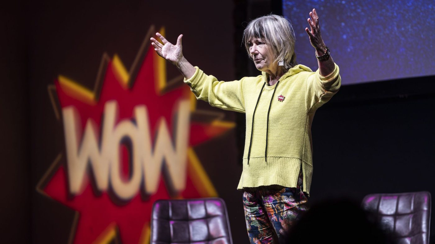 Jude (a white woman in a yellow top) stands on a stage with her hands in the air