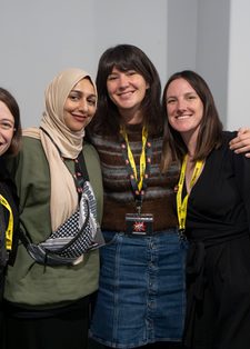 Four women smiling and posing closely together, wearing yellow lanyards, in a room with a neutral background. The mood is friendly and joyful.