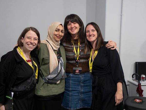 Four women smiling and posing closely together, wearing yellow lanyards, in a room with a neutral background. The mood is friendly and joyful.