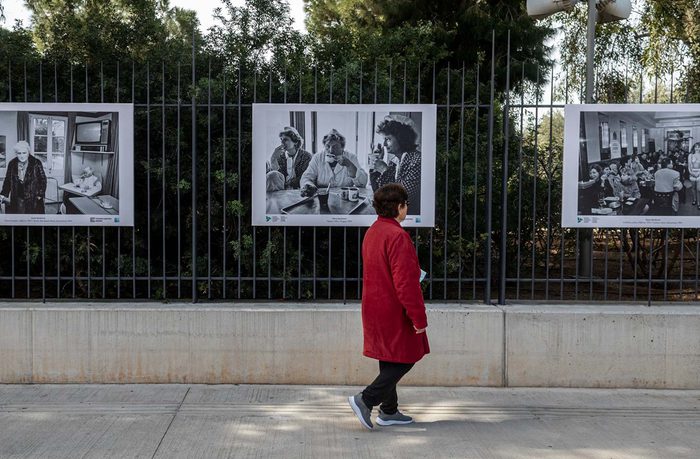 A woman in a red coat walks past a fence displaying three large black-and-white photos, set against a background of green foliage.