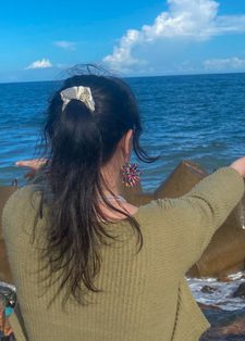 A woman with long dark hair in a ponytail, wearing a green sweater, faces the ocean. She gestures to the sea, surrounded by large rocks