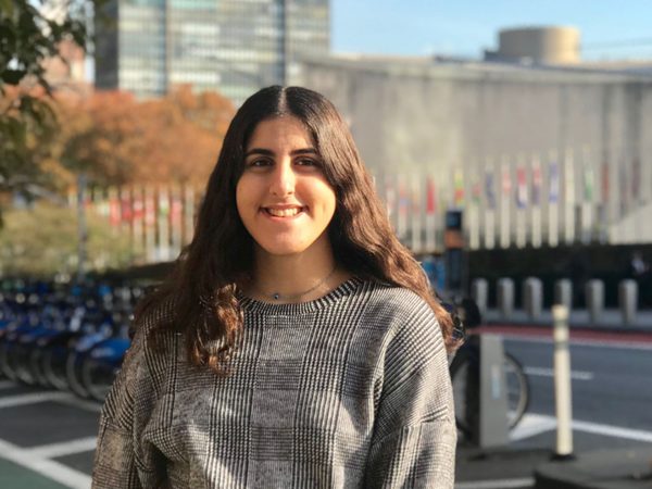 Rym Badran smiles at the camera with a bike rack behind her. She has long brown wavy hair and is wearing a grey check top.