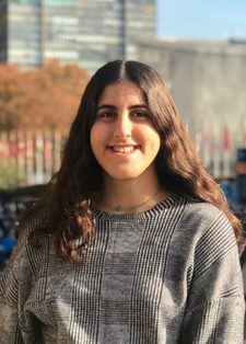 Rym Badran smiles at the camera with a bike rack behind her. She has long brown wavy hair and is wearing a grey check top.