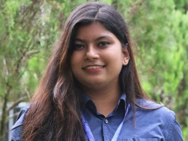Saiban Binte Shahaj looks into the camera with trees behind her. She has long brown hair and is wearing a blue shirt and lanyard.