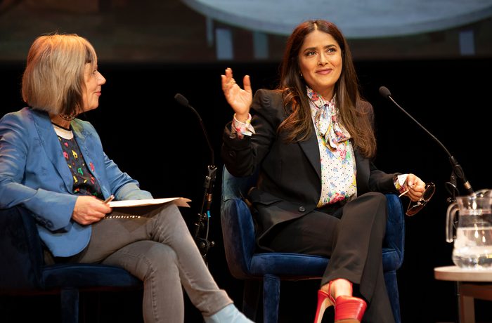 Salma Hayek and Jude Kelly sit on stage in conversation. Jude wears a blue jacket with a clipboard and Salma wears a black suit and floral blouse, gesturing expressively.