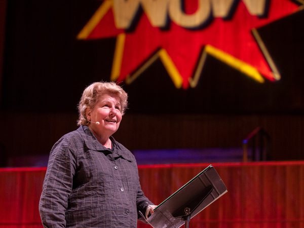 Sandi Toksvig smiling confidently on stage with a large red and yellow "WOW" sign projected behind them