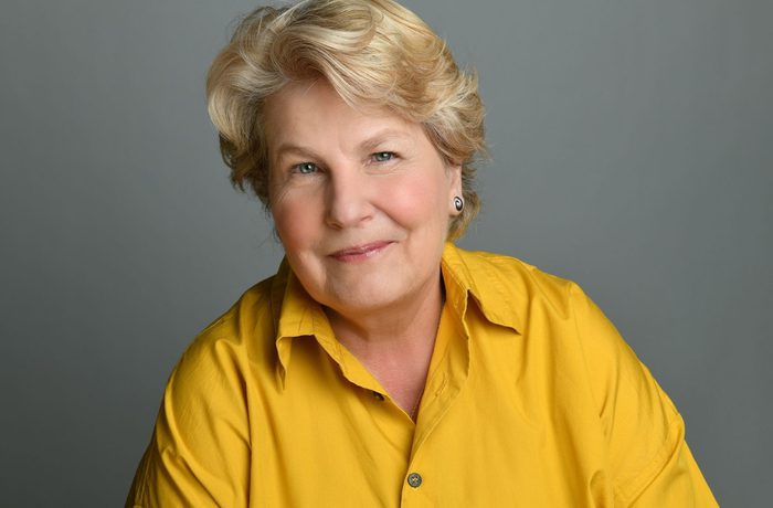 Sandi Toksvig looks into the camera with a slight smile. She has short blonde hair and is wearing a yellow shirt.