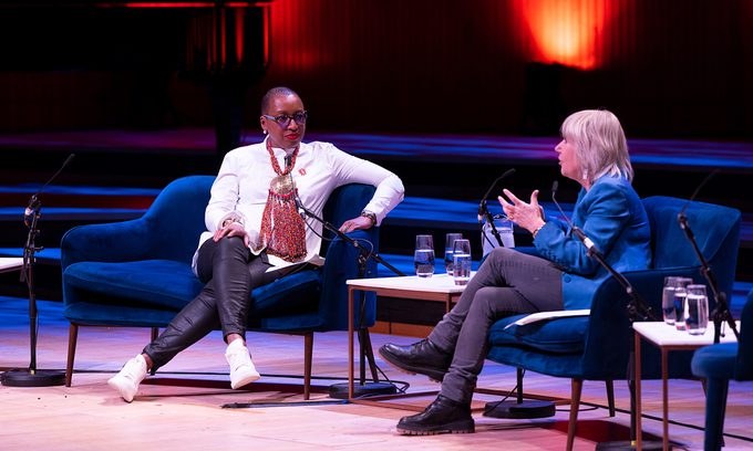 Two people are seated on stage in a discussion. Both sit on blue armchairs with microphones and water glasses on tables.
