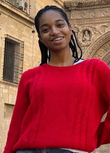 A person in a red sweater smiles warmly in front of a stone building with an arched entrance