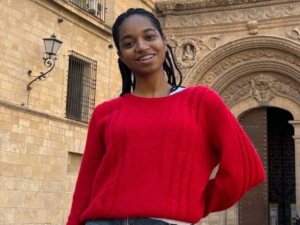 A person in a red sweater smiles warmly in front of a stone building with an arched entrance