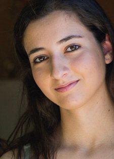 Young woman with long dark hair, smiling gently, leans against a wooden wall.