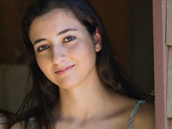 Young woman with long dark hair, smiling gently, leans against a wooden wall.